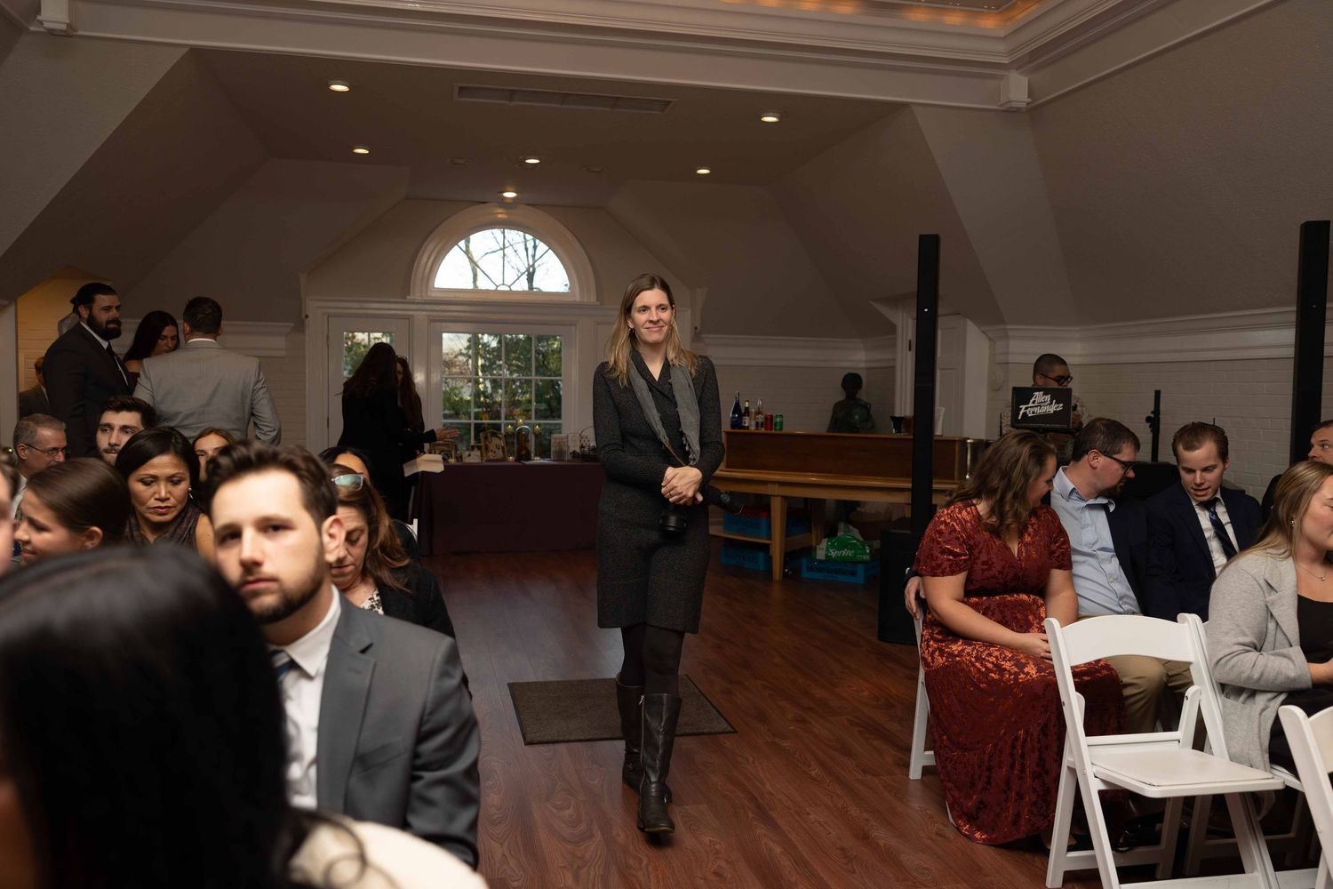 People gather in an elegant indoor venue with arched windows and white chairs during what appears to be a formal event.