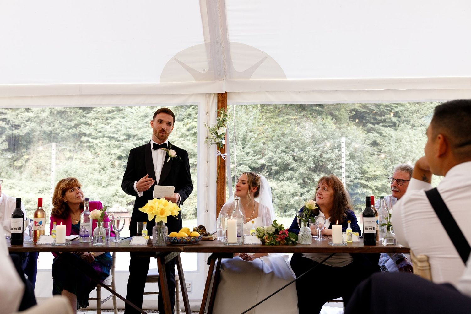 Wedding guests seated at tables under a white tent raise their glasses in a toast during an outdoor reception celebration.