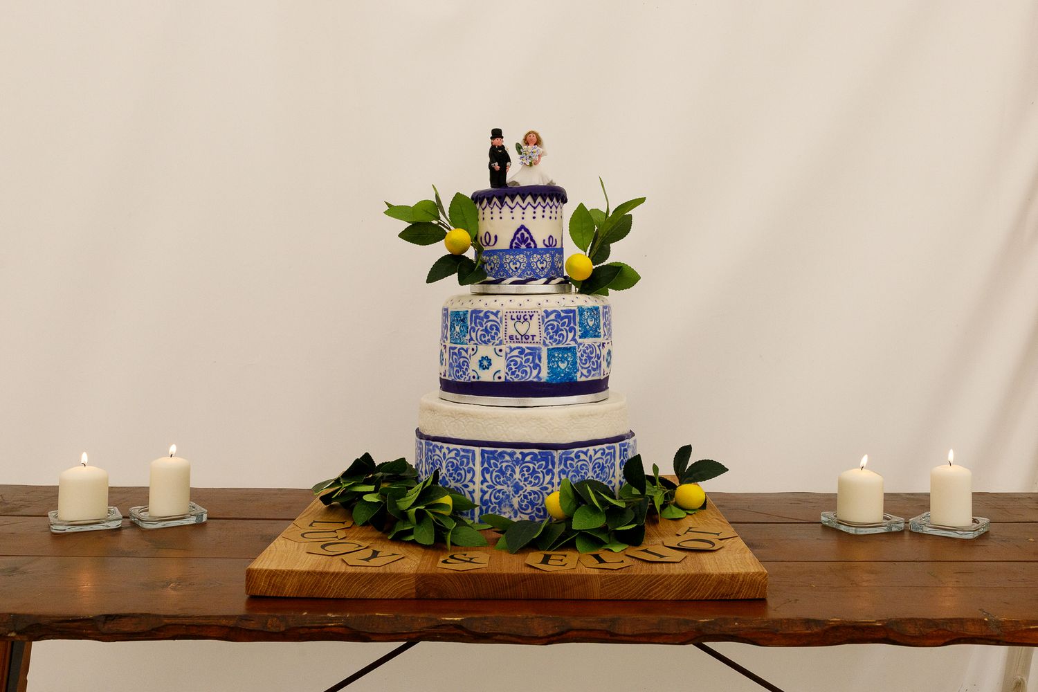 Elegant white wedding cake with blue and purple floral accents displayed on a wooden table with greenery and candles.