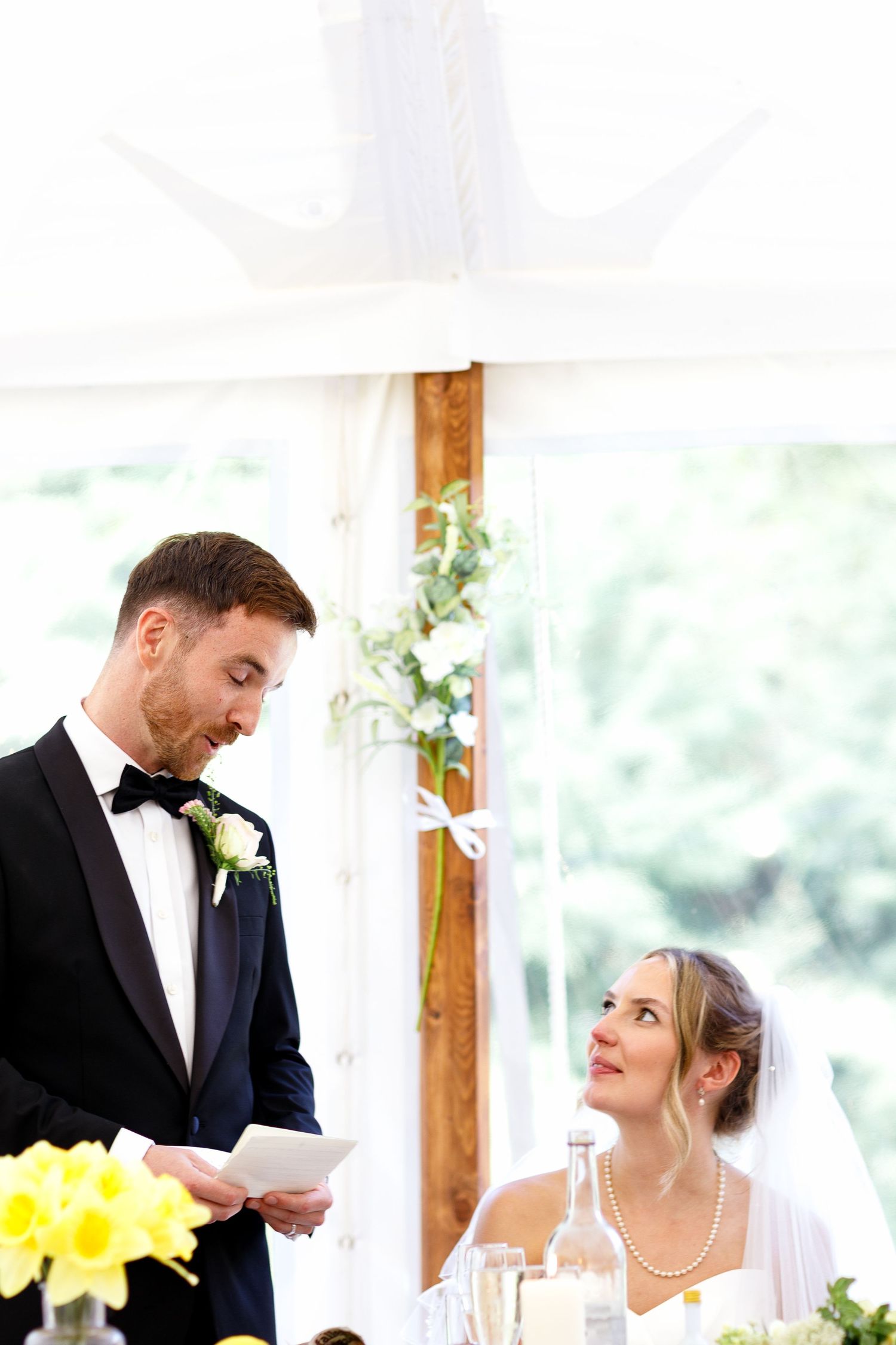 Bride and groom share a candid moment during their wedding reception at a light-filled venue with yellow flowers.