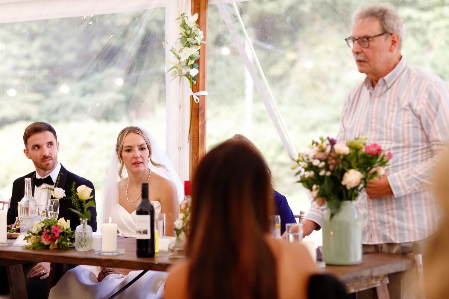 A candid moment during a wedding reception shows guests seated at tables under a white tent with floral centerpieces.