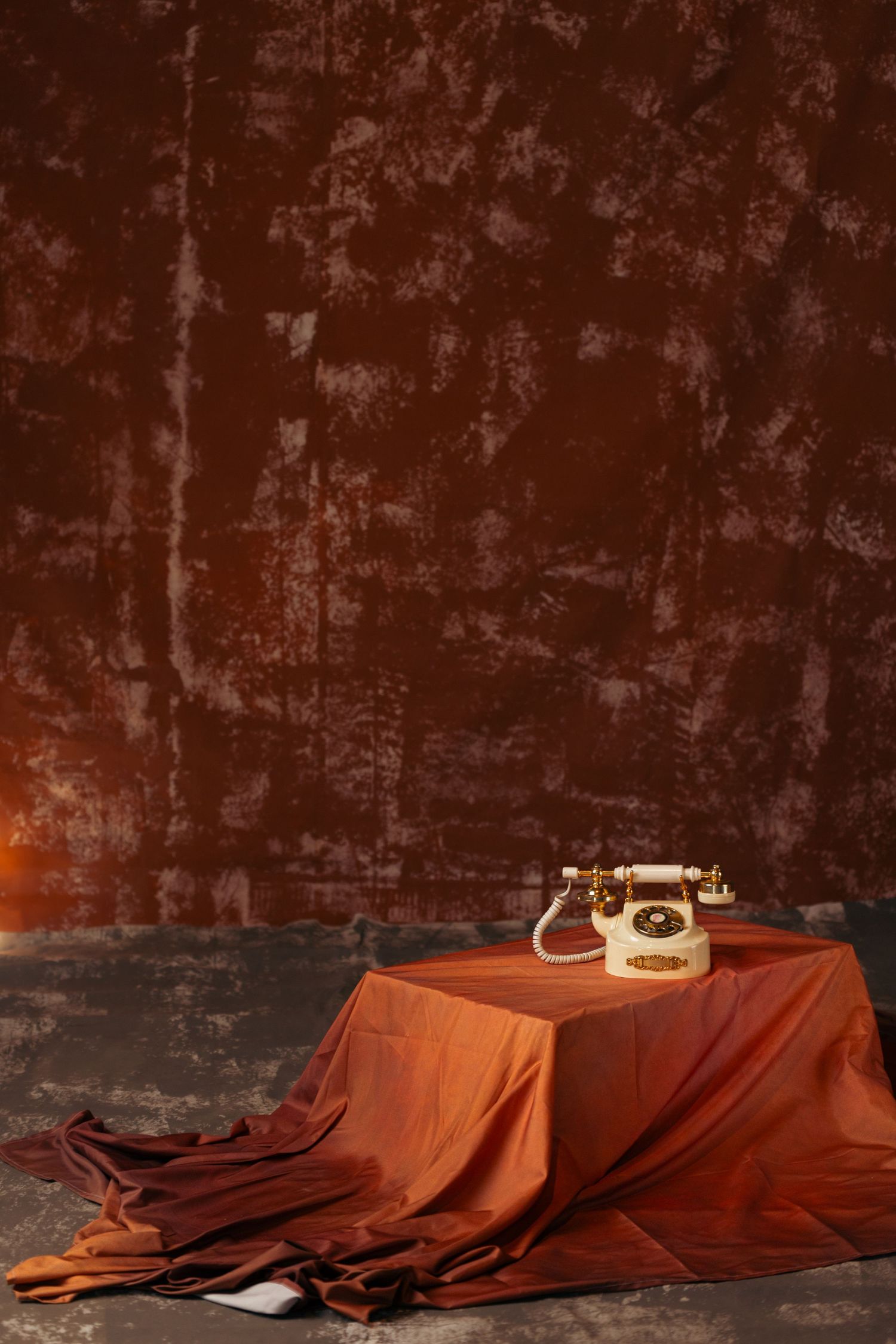 A vintage rotary telephone sits on a brown cloth draped surface against a dark textured wall.