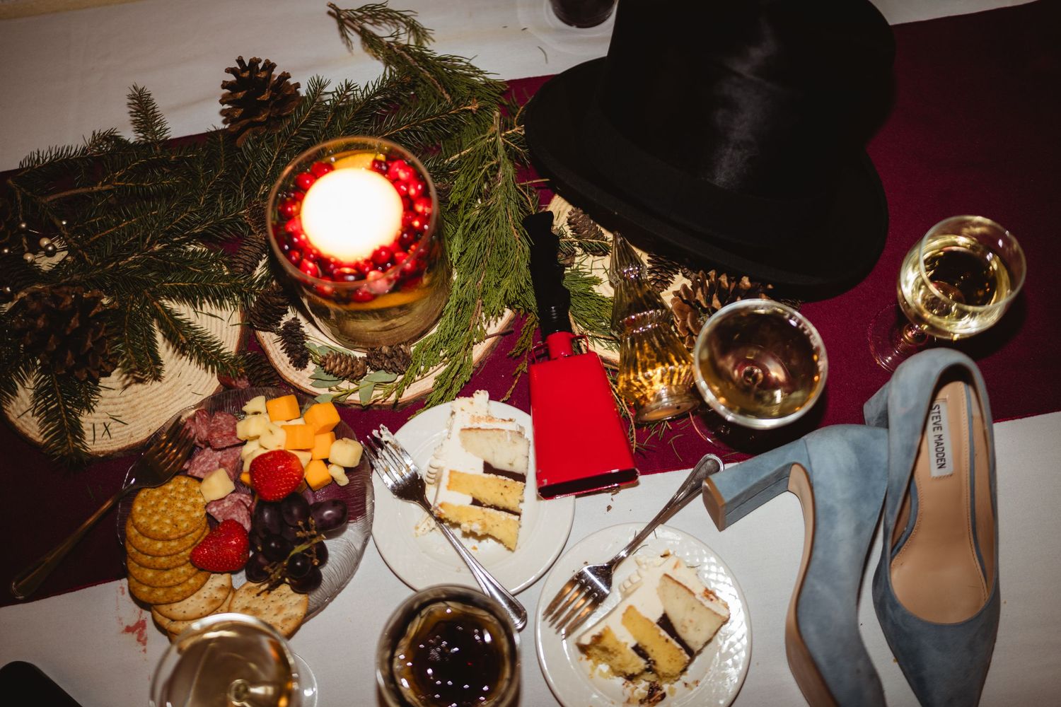 Overhead view of a holiday dinner table with candles, wine glasses, shoes, and appetizer plates on a red tablecloth.