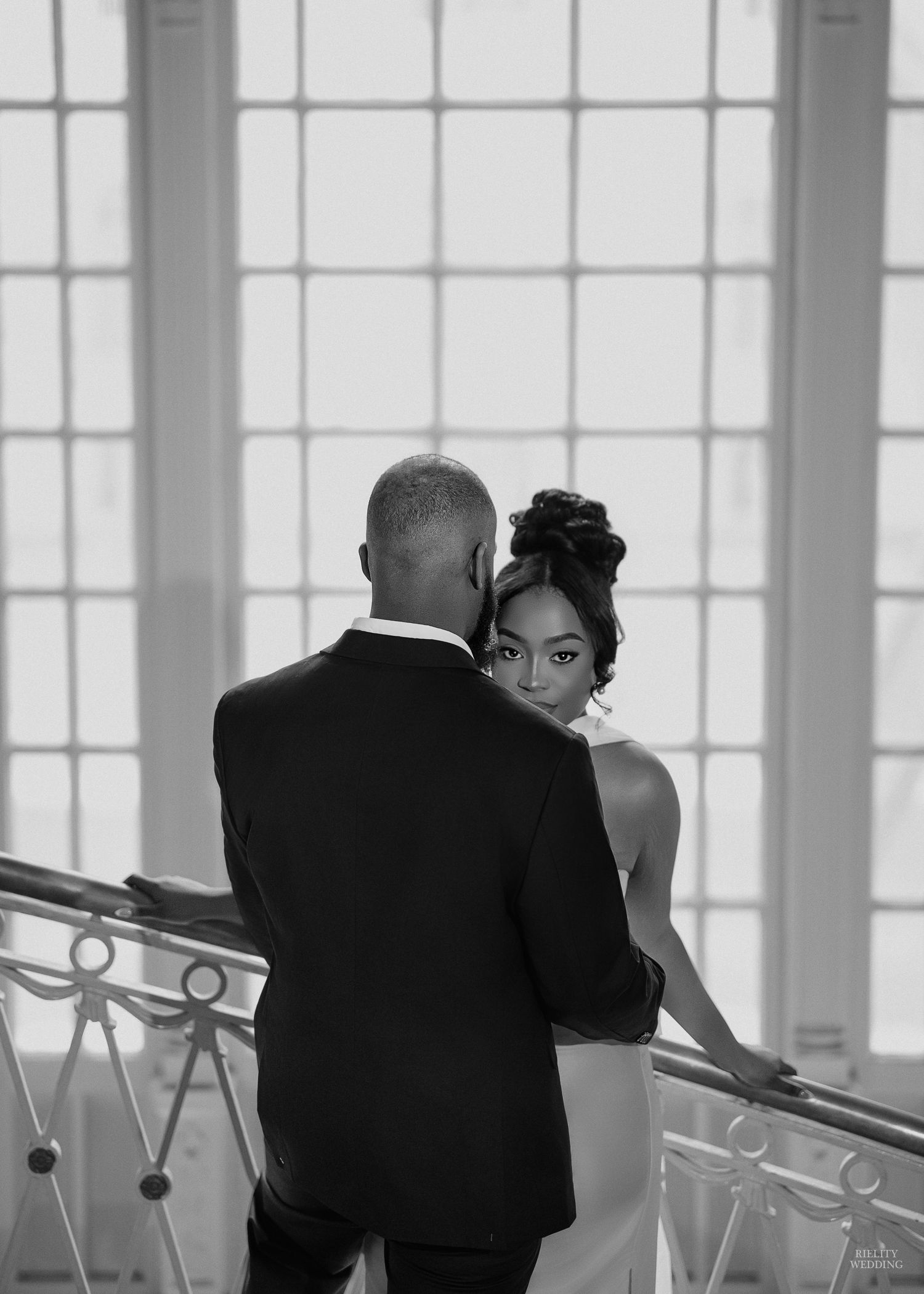 A couple in formal attire shares an intimate dance in an elegant room with large windows.