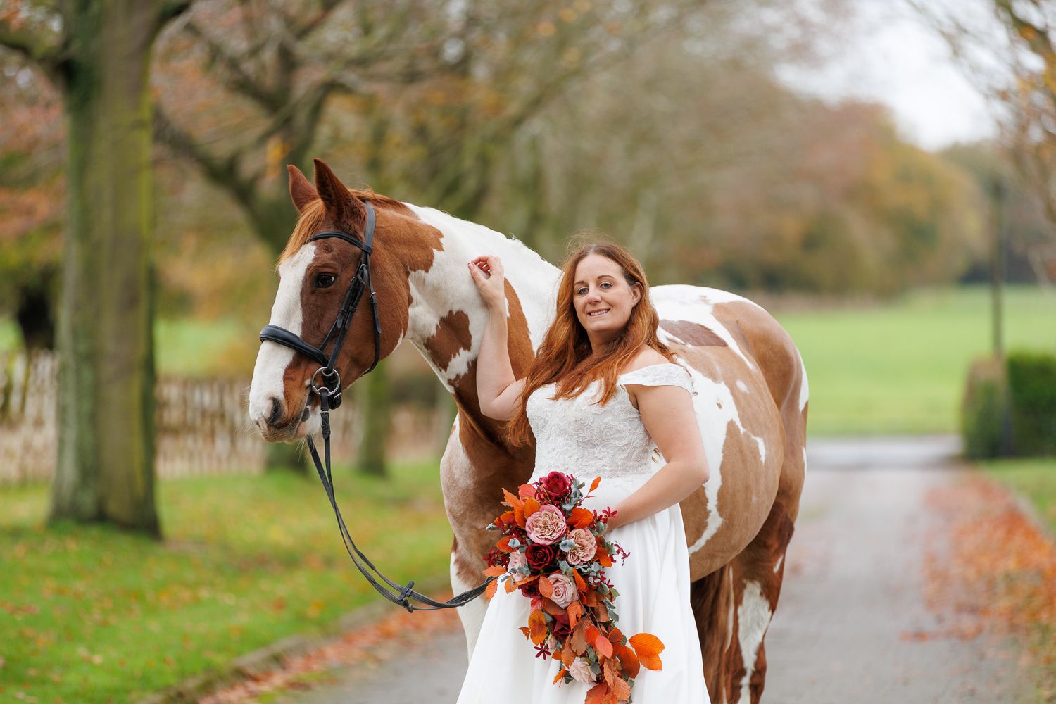 A Paint horse stands on a tree-lined path covered in fallen autumn leaves during a fall photo session.