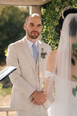 Groom in light beige suit and blue tie standing under floral-decorated wedding arch.