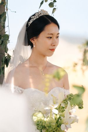 Elegant bride in white dress poses with delicate white flowers and greenery in soft natural light.