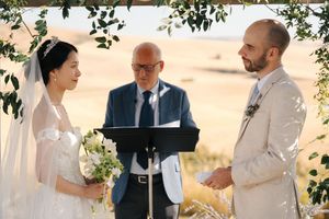 Outdoor wedding ceremony setup with greenery arch and guests in a scenic field setting.