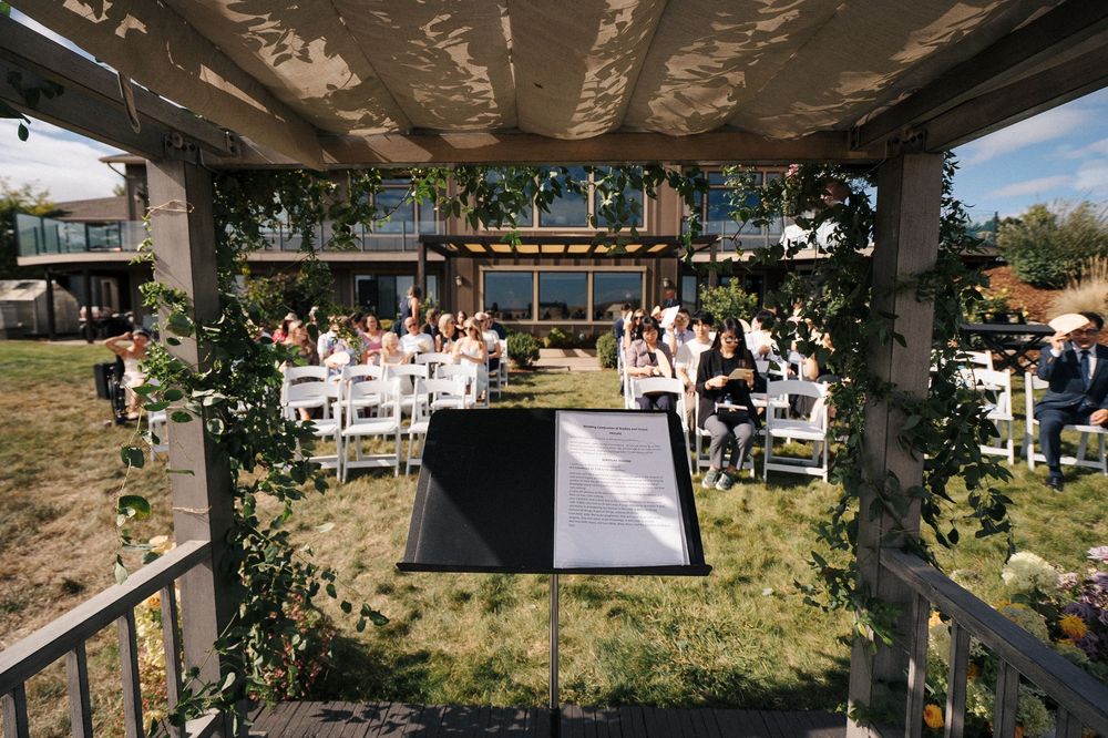 Outdoor wedding ceremony setup under a pergola with white chairs arranged on a grassy lawn.