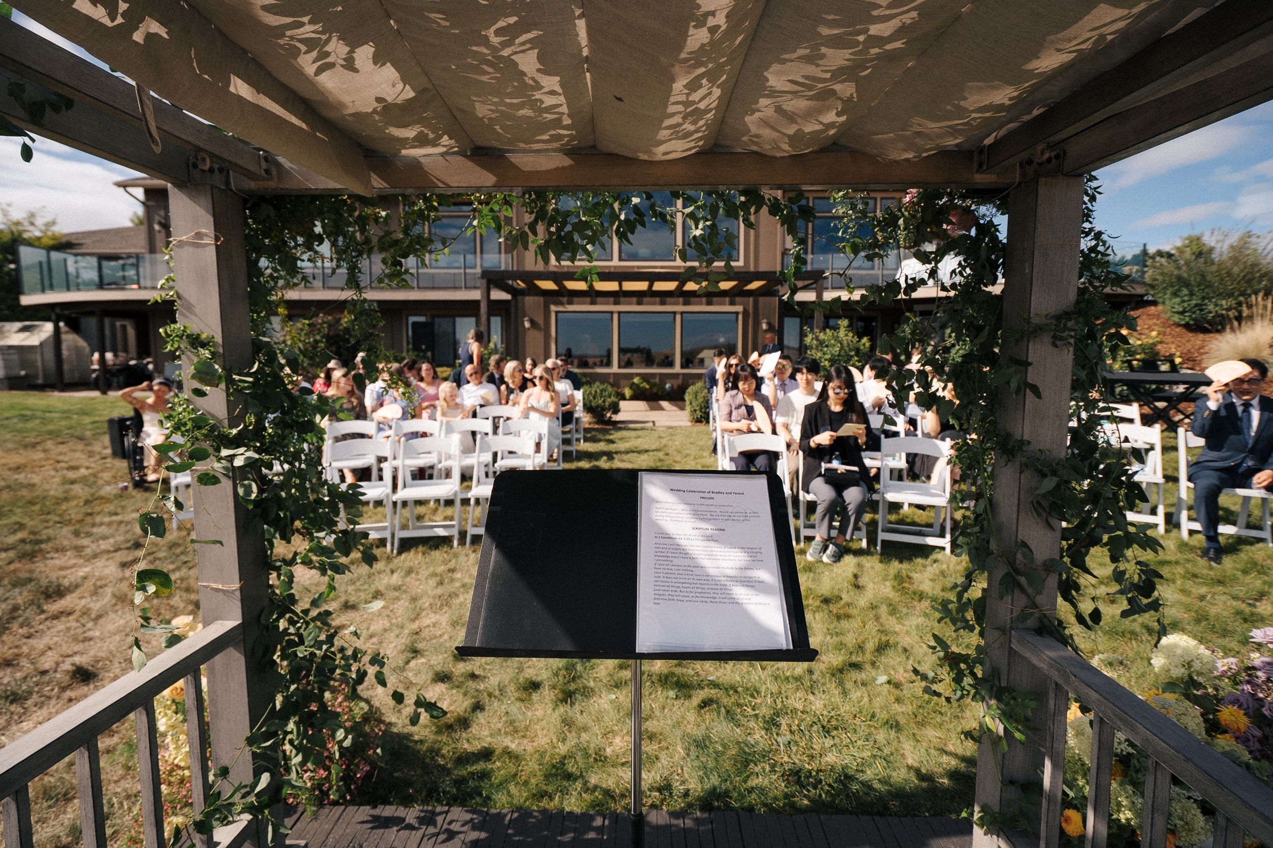 Outdoor wedding ceremony setup under a pergola with white chairs arranged on a grassy lawn.