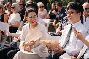 Wedding guests seated outdoors smile and use handheld fans on a sunny day.