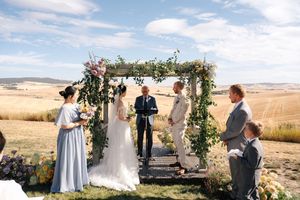 Outdoor wedding ceremony with guests gathered under decorated arch in rural hillside setting.