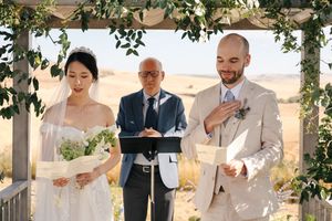 Outdoor wedding ceremony with guests seated under leafy green arbor in rustic setting.