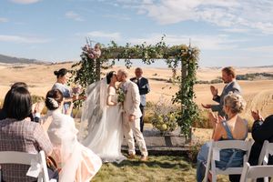 Wedding ceremony guests gather outdoors under a floral arch with scenic mountain views in the background.