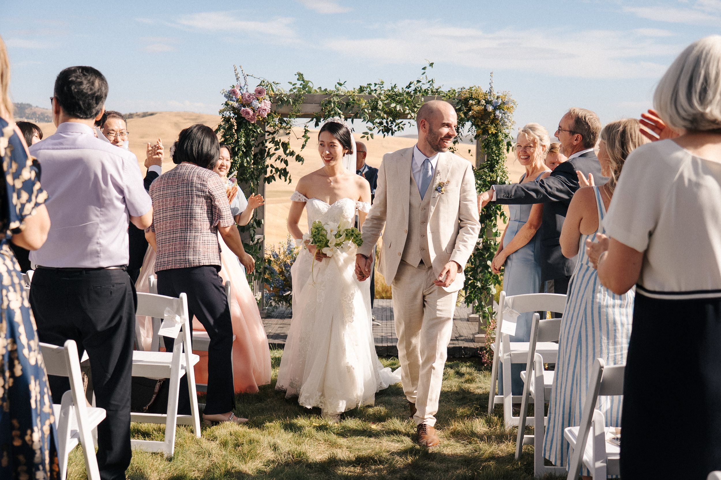 Guests stand in white attire at an outdoor wedding ceremony with floral decorations and white chairs.