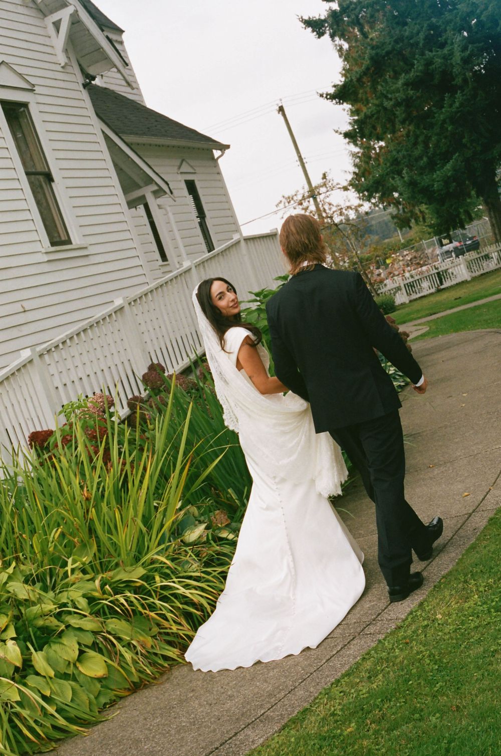A bride in a white wedding dress and groom in black suit walk along a path beside a white church building.