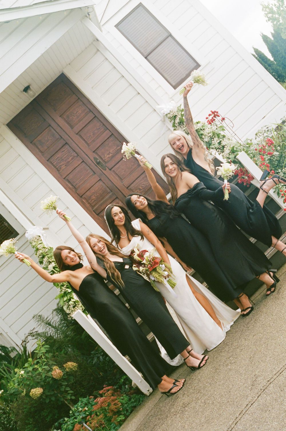 Wedding party members in formal attire pose together outside a white building with blooming flowers.