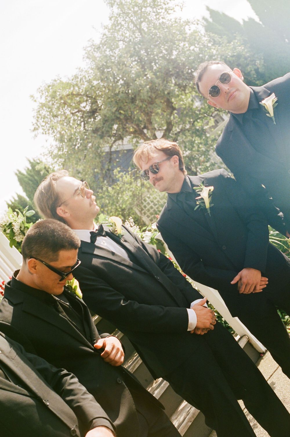 A sequence of candid shots showing groomsmen laughing together outdoors in black suits with sunlight filtering through trees.
