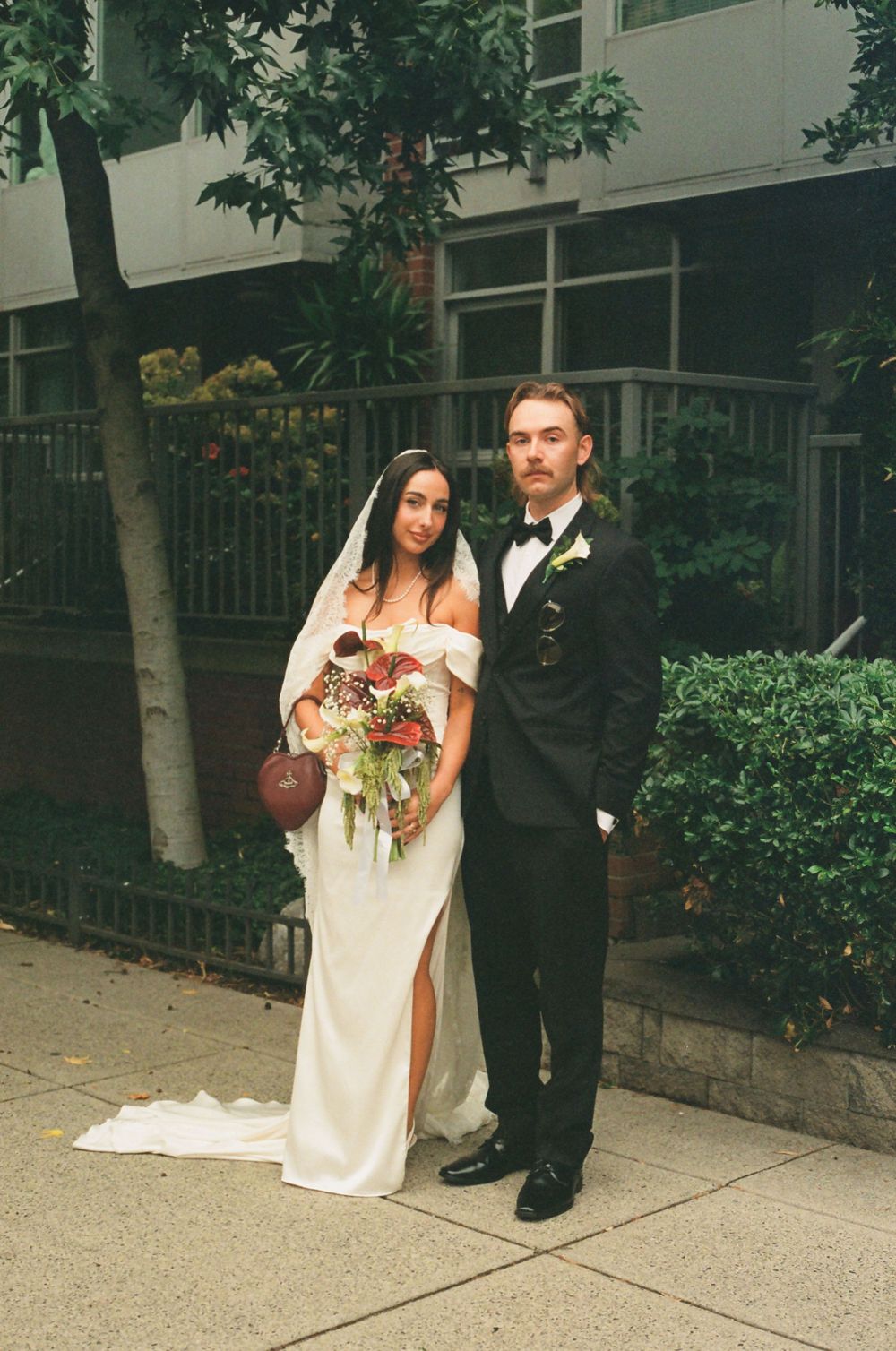 A couple poses outdoors on their wedding day, with the bride in a white gown holding a bouquet and groom in classic tuxedo.