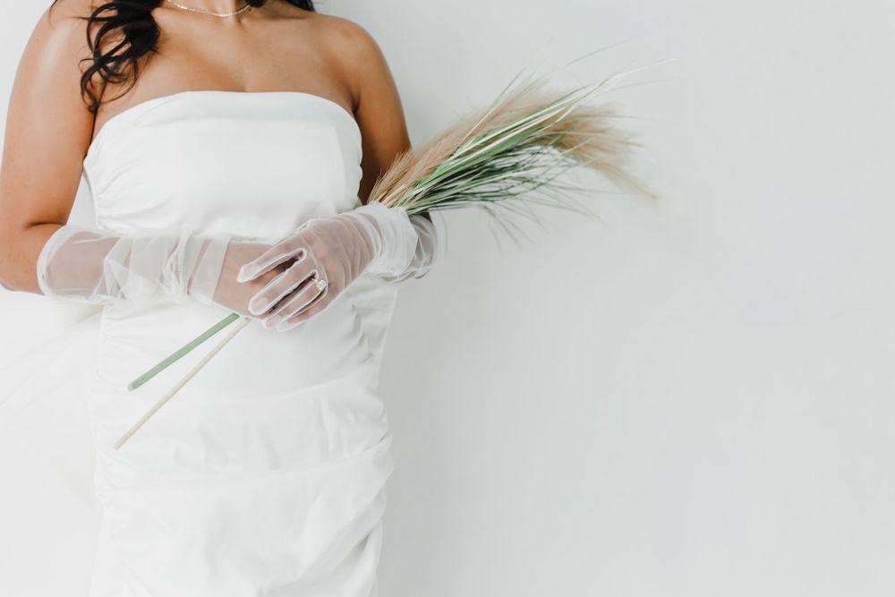 A person wearing white gloves gently holds a delicate green palm frond against a light gray background.