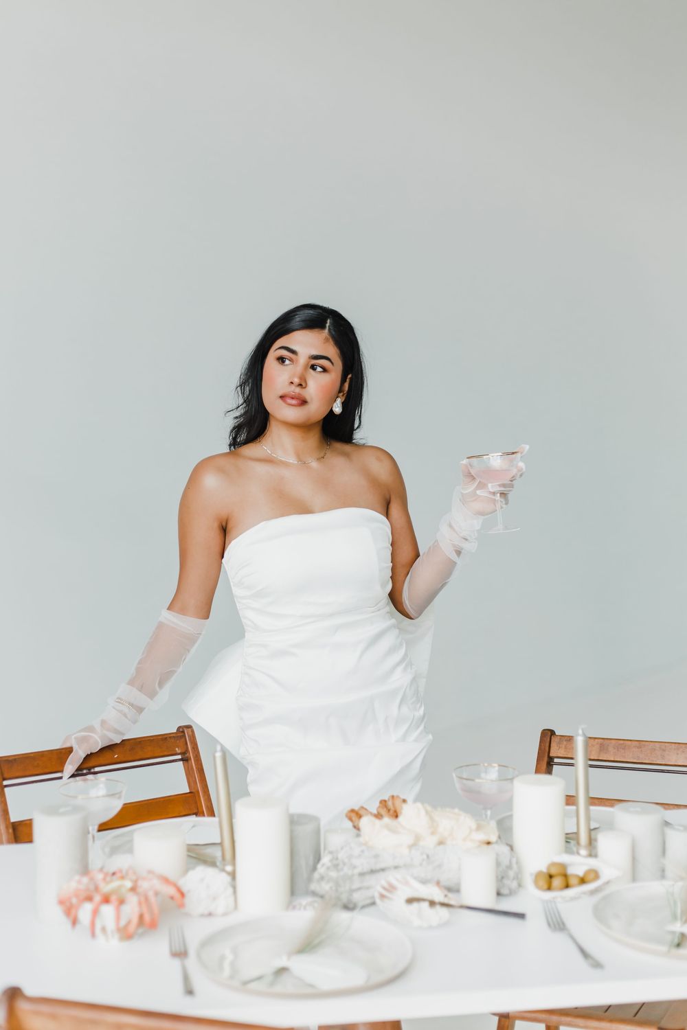Woman in white dress sits at elegant table setting with glassware and white decor against light gray backdrop.