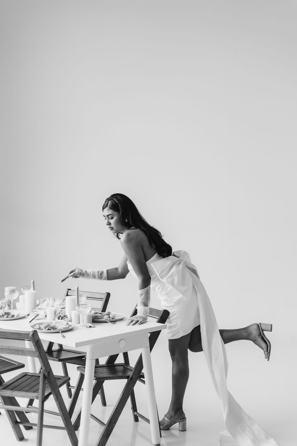 Person in white dress leans over elegant table setting in black and white photo.