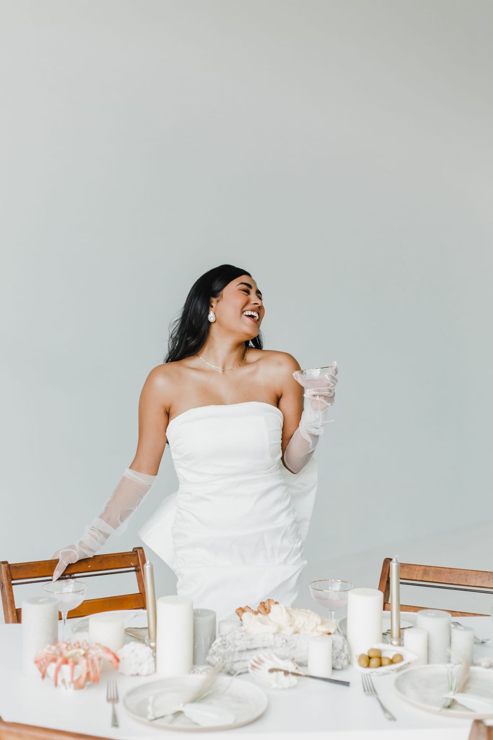 A person in a white dress laughs joyfully at an elegant table setting with desserts and drinks.