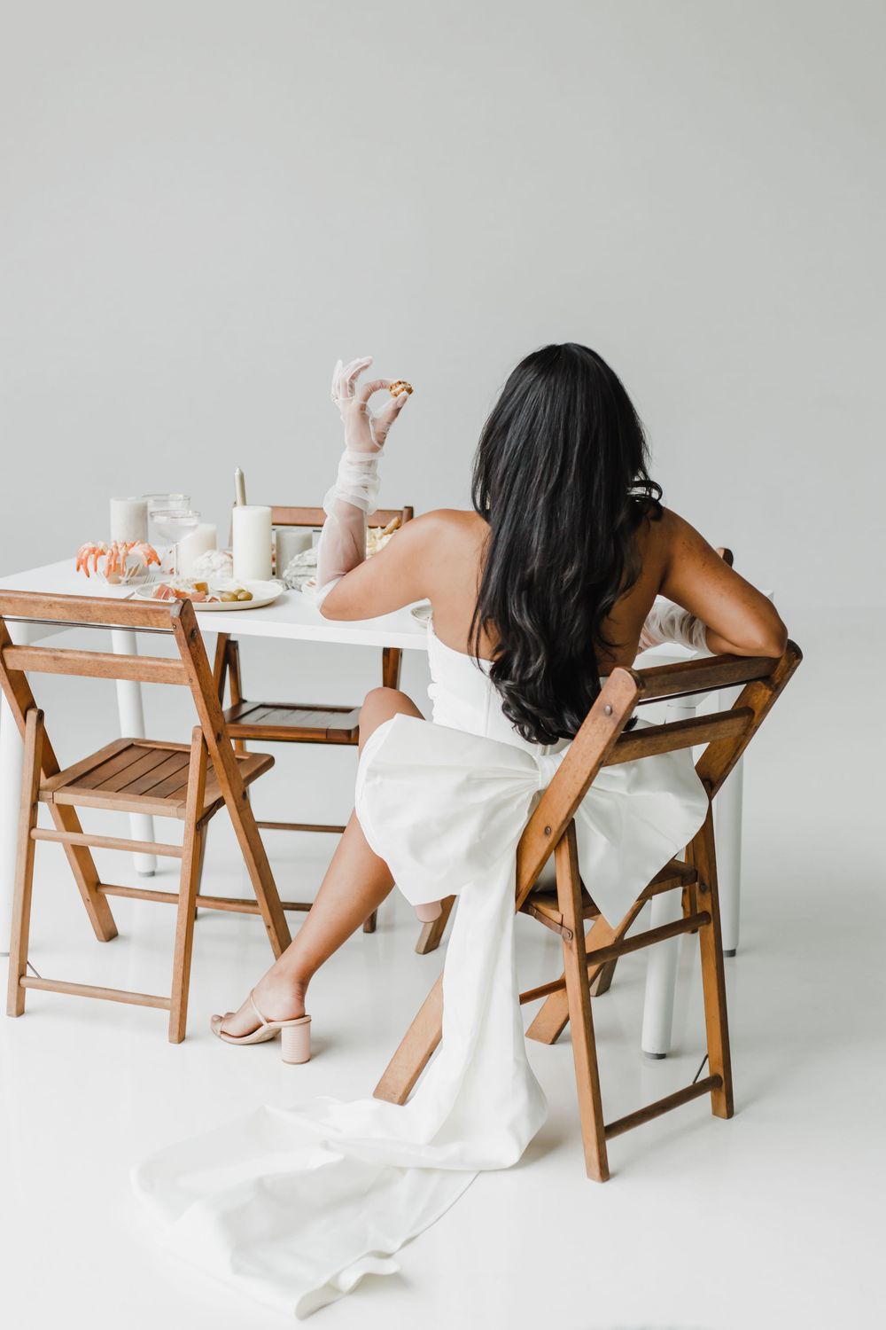 Person in white dress sits at table with refreshments against minimalist background.
