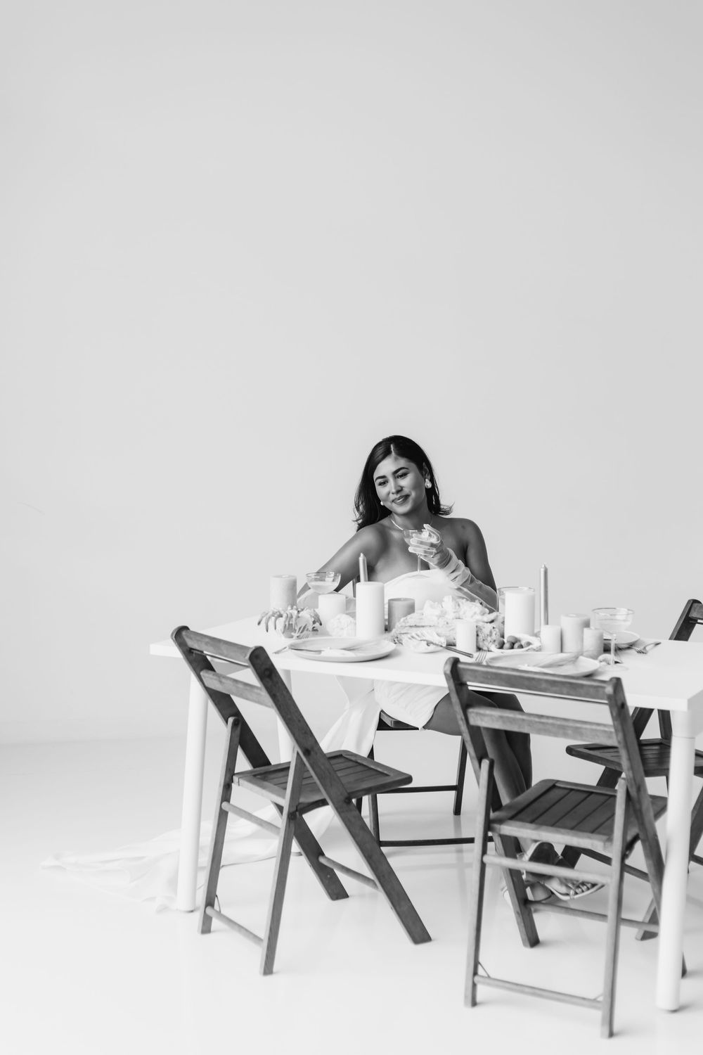 A person in a white dress sits at an elegant table setting with wooden chairs against a light background.