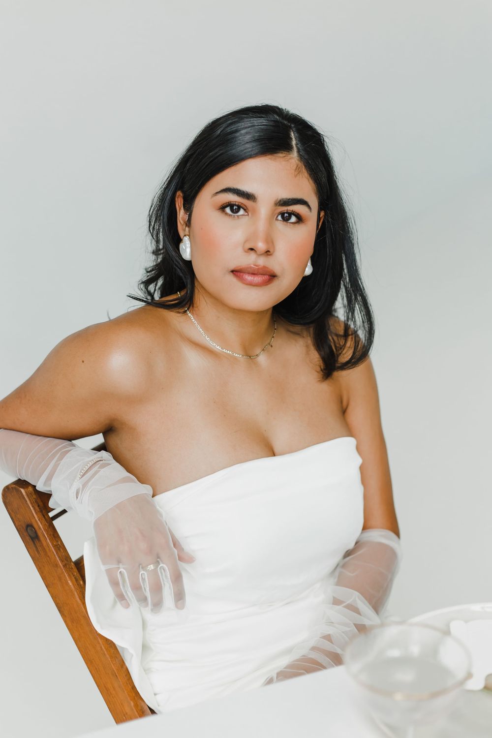 Woman in white strapless dress and sheer gloves poses elegantly against light backdrop.