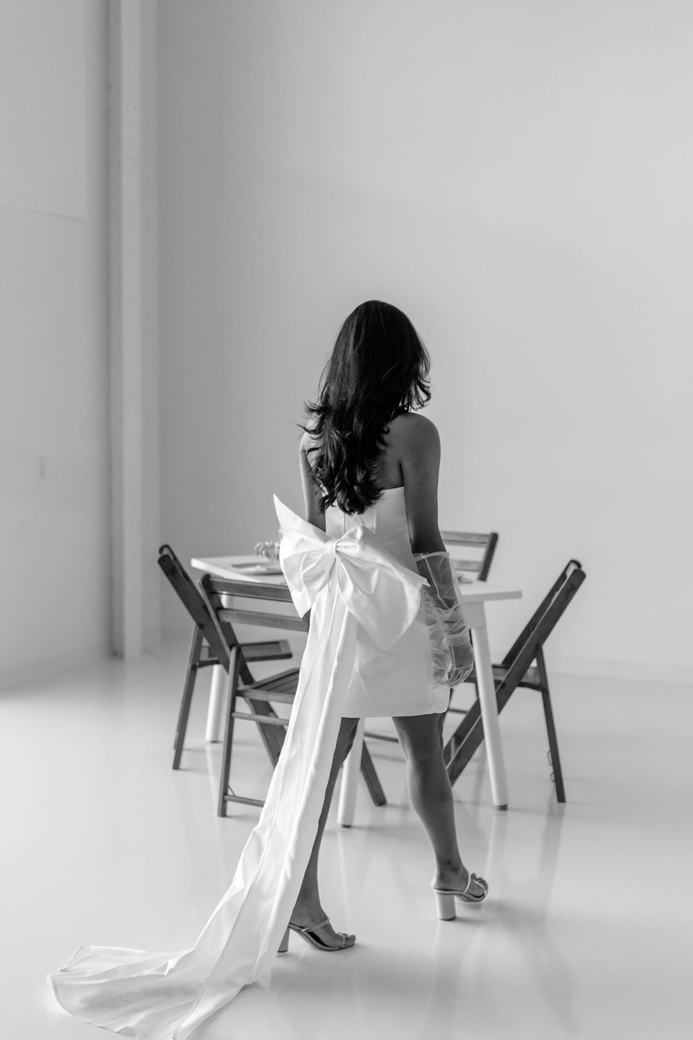 A figure in a white dress sits gracefully on a wooden chair in a minimalist white room setting.