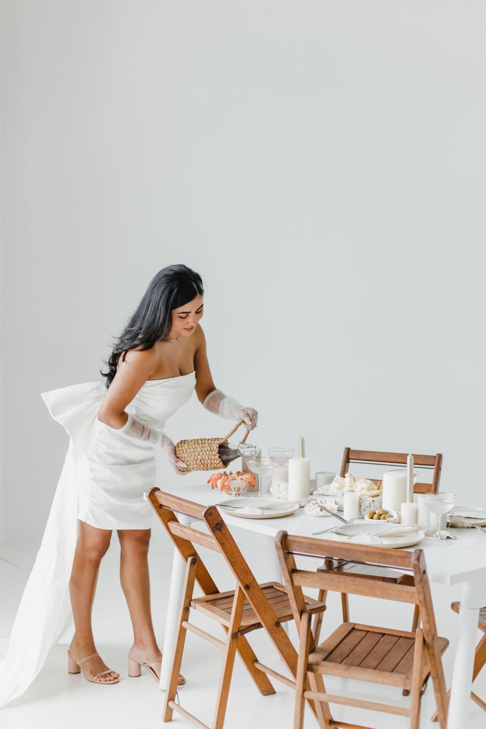 A white-dressed figure interacts with a table setting featuring wooden chairs in a bright space.