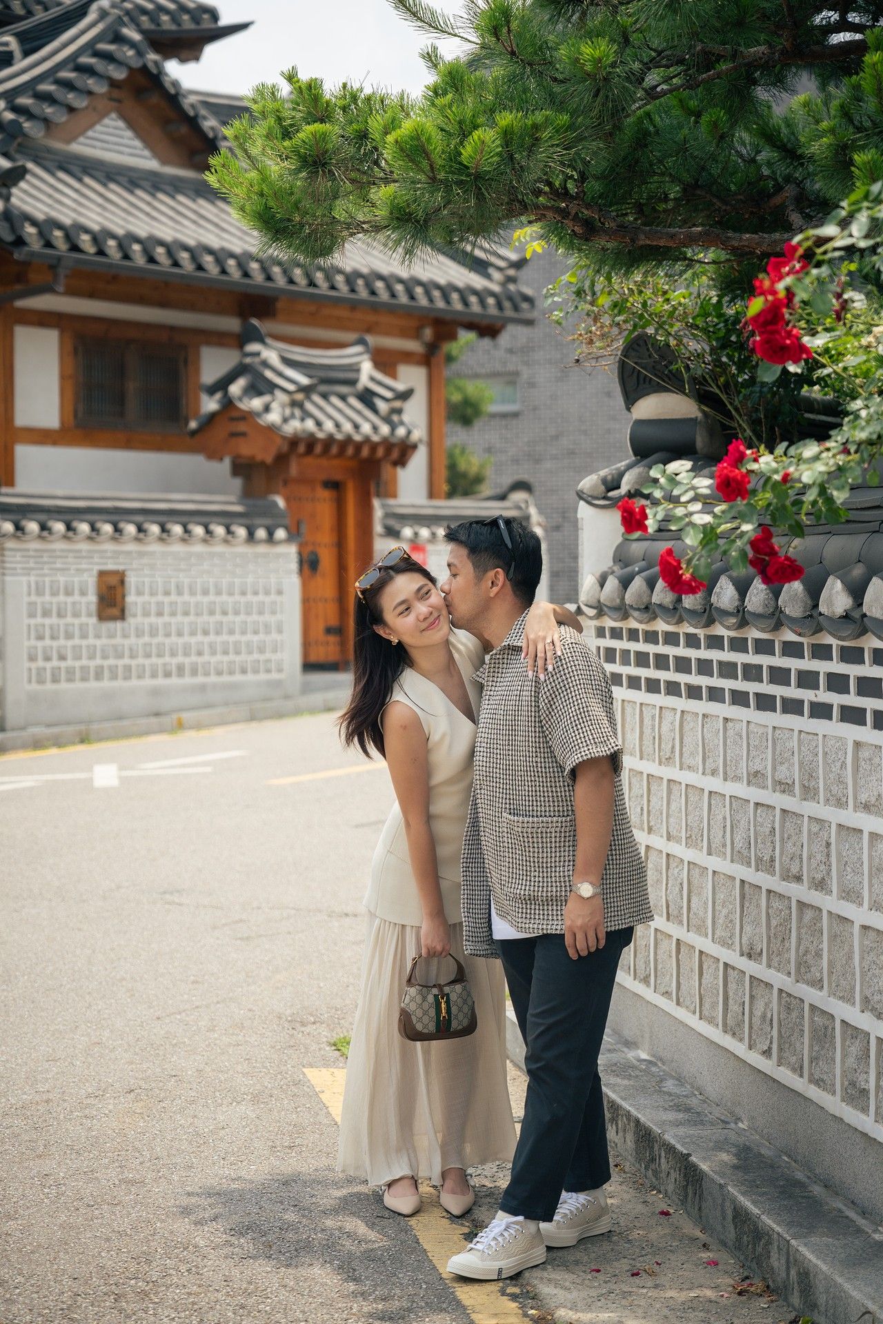 A couple shares a romantic moment in front of traditional Korean architecture with white walls and tiled roofs.