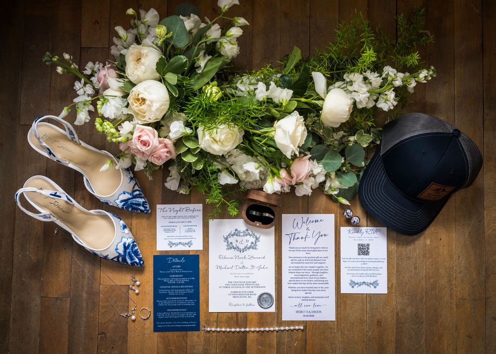Wedding details flatlay with white floral arrangement, invitations, shoes and accessories on wooden surface.