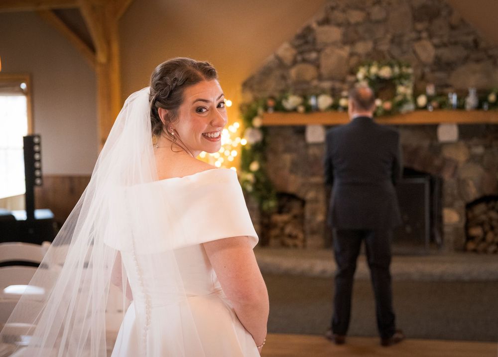 Bride in classic white dress looks over shoulder with stone fireplace and warm lighting in background.