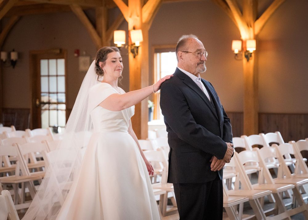 Wedding first look moment captured in rustic venue with wooden beams and white chairs.