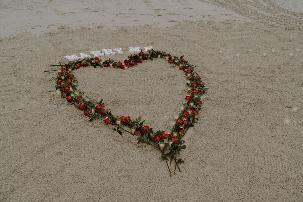 Red rose petals and greenery forming a heart design on beach sand.