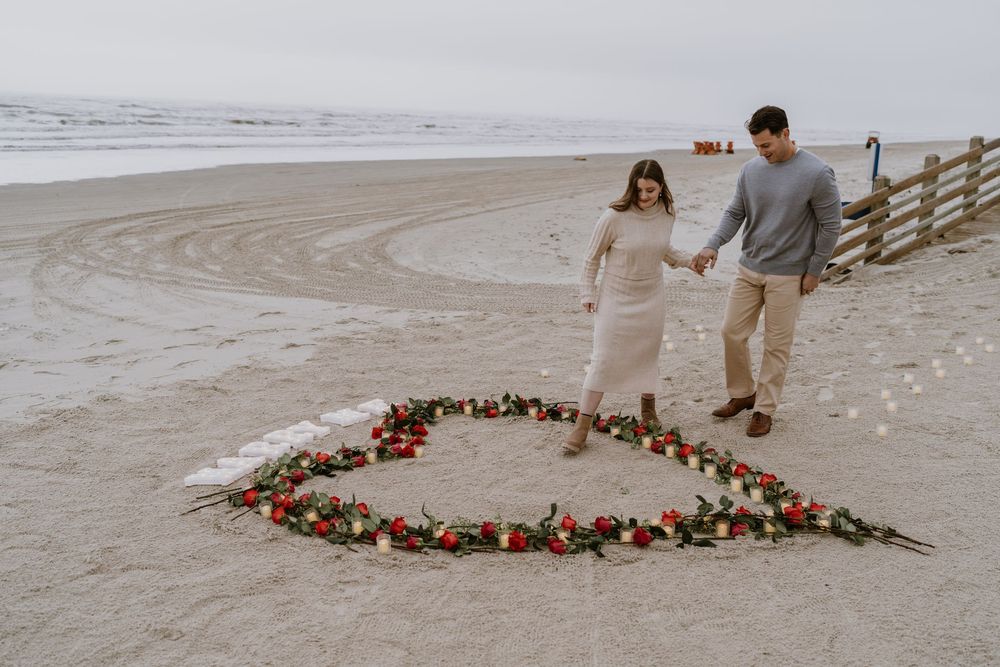 A couple walks along beach near heart-shaped floral arrangement on sand.