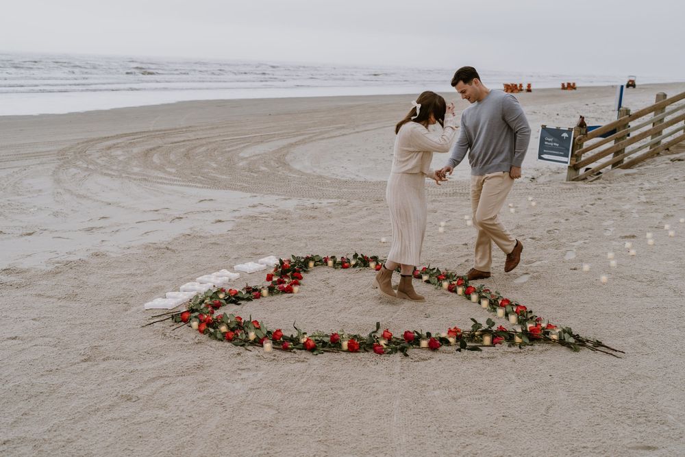A couple stands inside a heart shape made of roses on a misty beach.