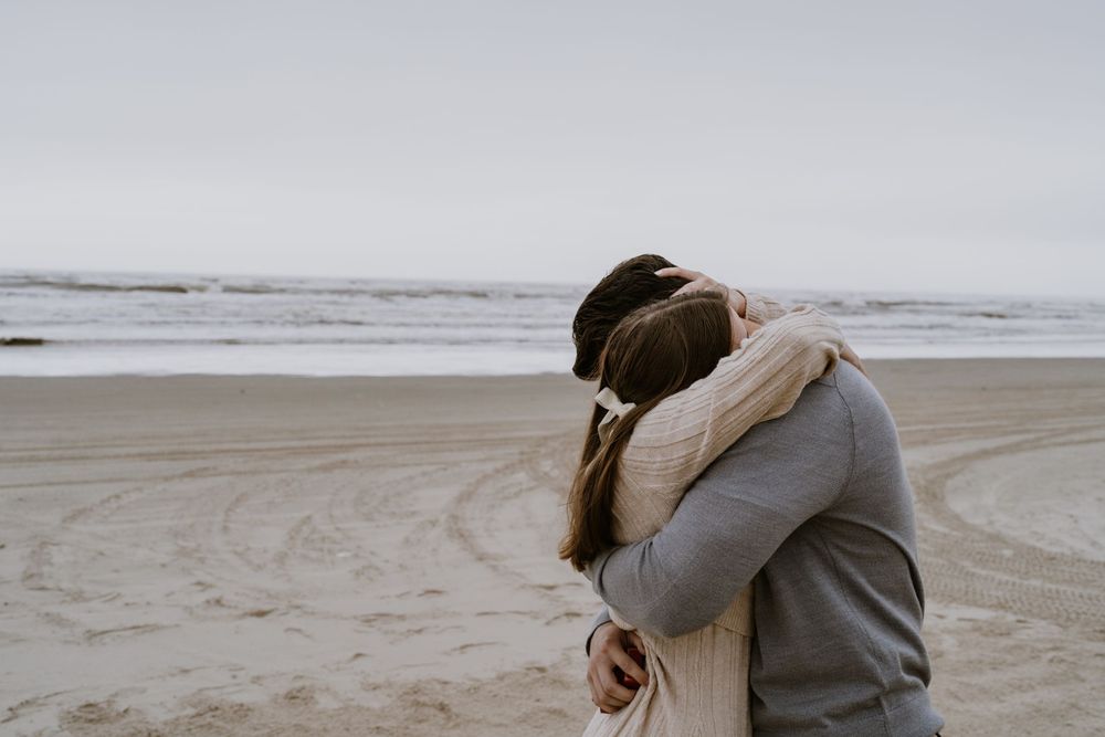 A couple embraces on a cloudy beach after a proposal.