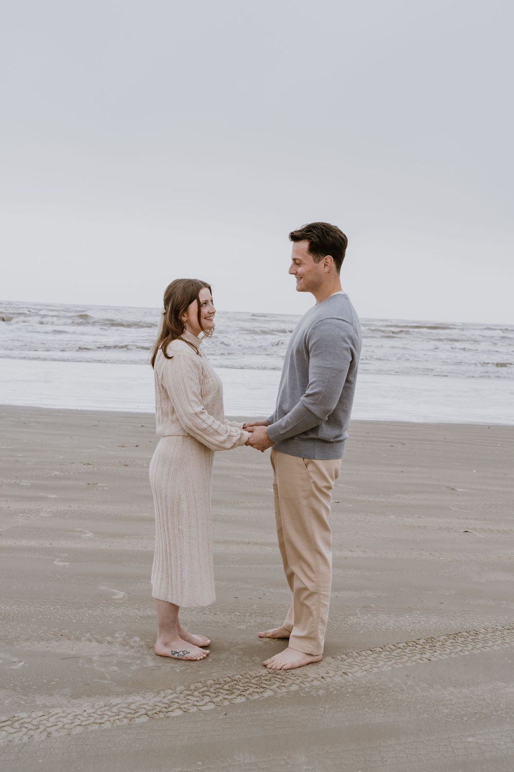 A couple stands face to face holding hands on a misty beach.