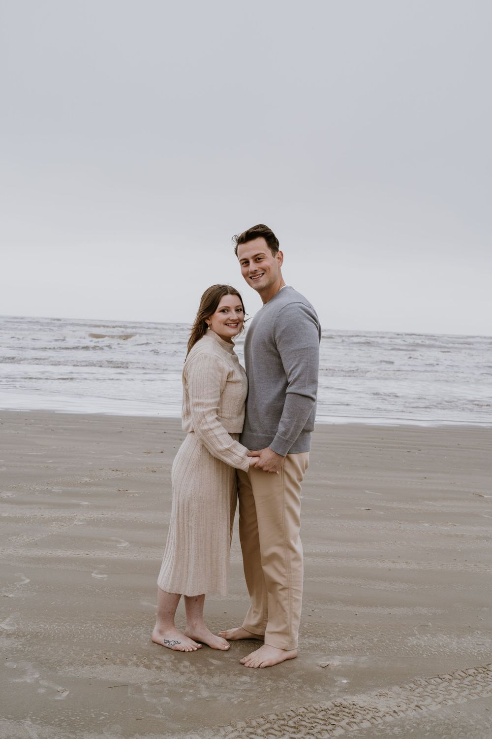 A newly engaged couple poses together on a cloudy beach.