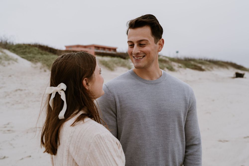 A couple smiles at each other while standing on the beach.