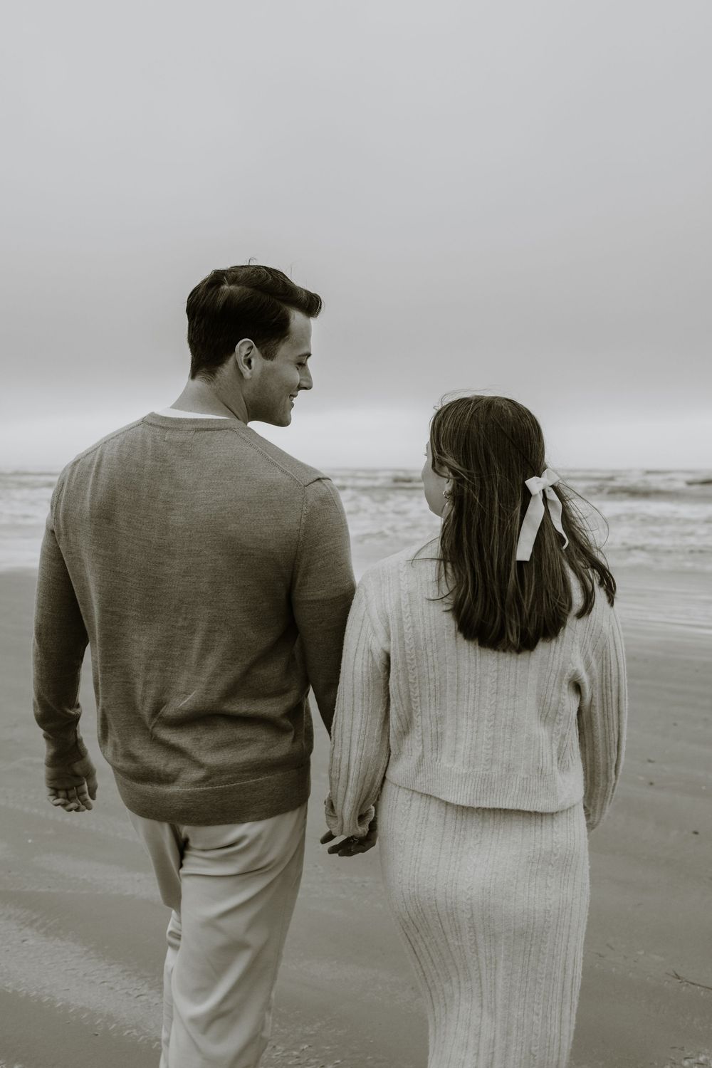 A couple walks hand in hand along a moody black and white beach.