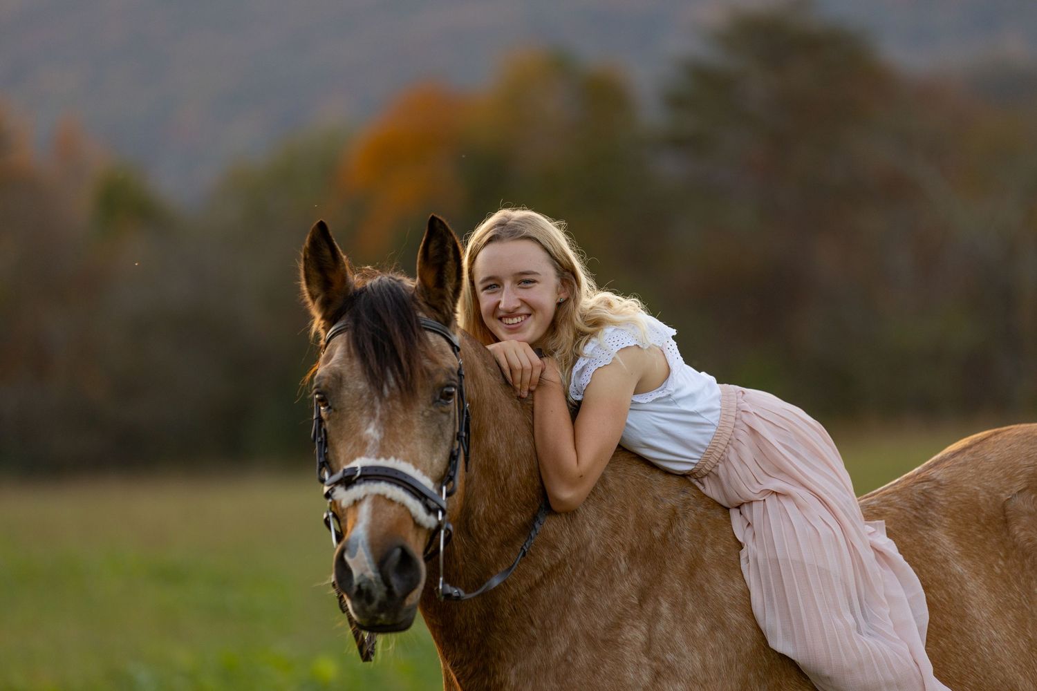 A young person in a pink dress rides a brown horse through a scenic outdoor setting at sunset.