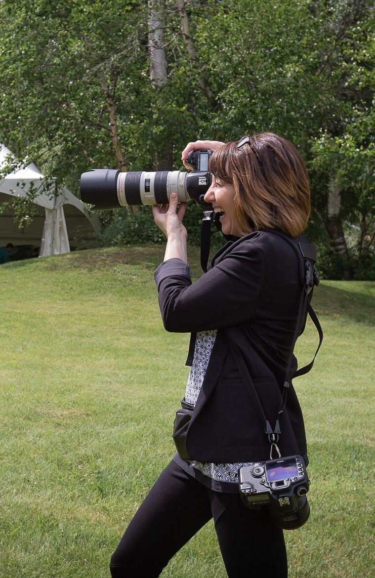 A photographer in black clothing uses a telephoto lens to capture shots in a green outdoor setting.