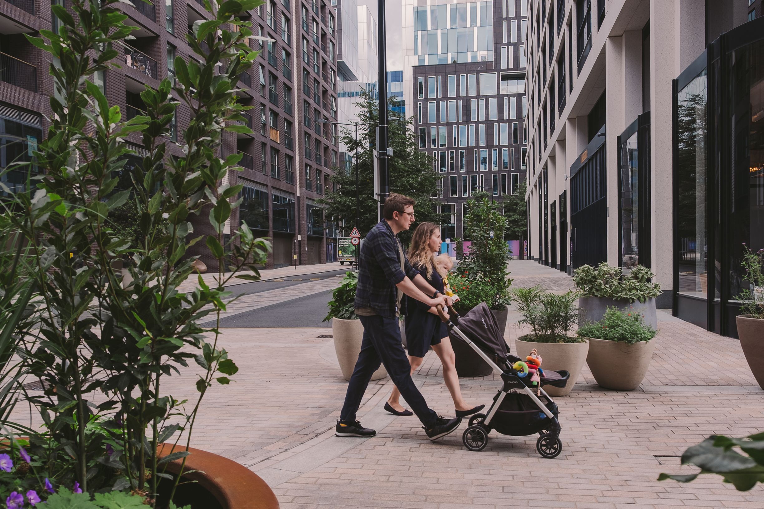 Modern urban sidewalk with tall buildings, planters, and people walking in the background.