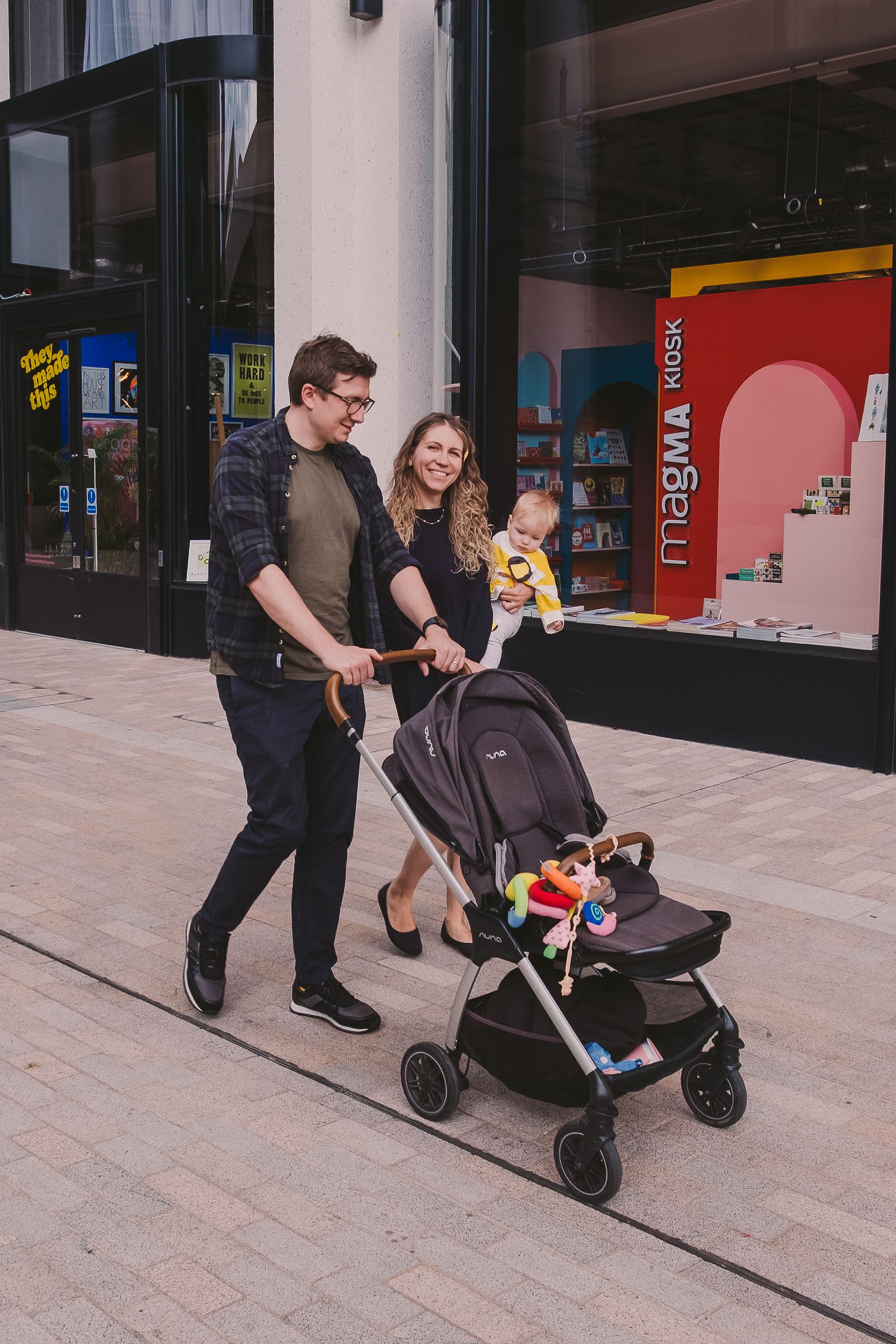 People walking along a shopping area with storefronts and pushing a stroller.