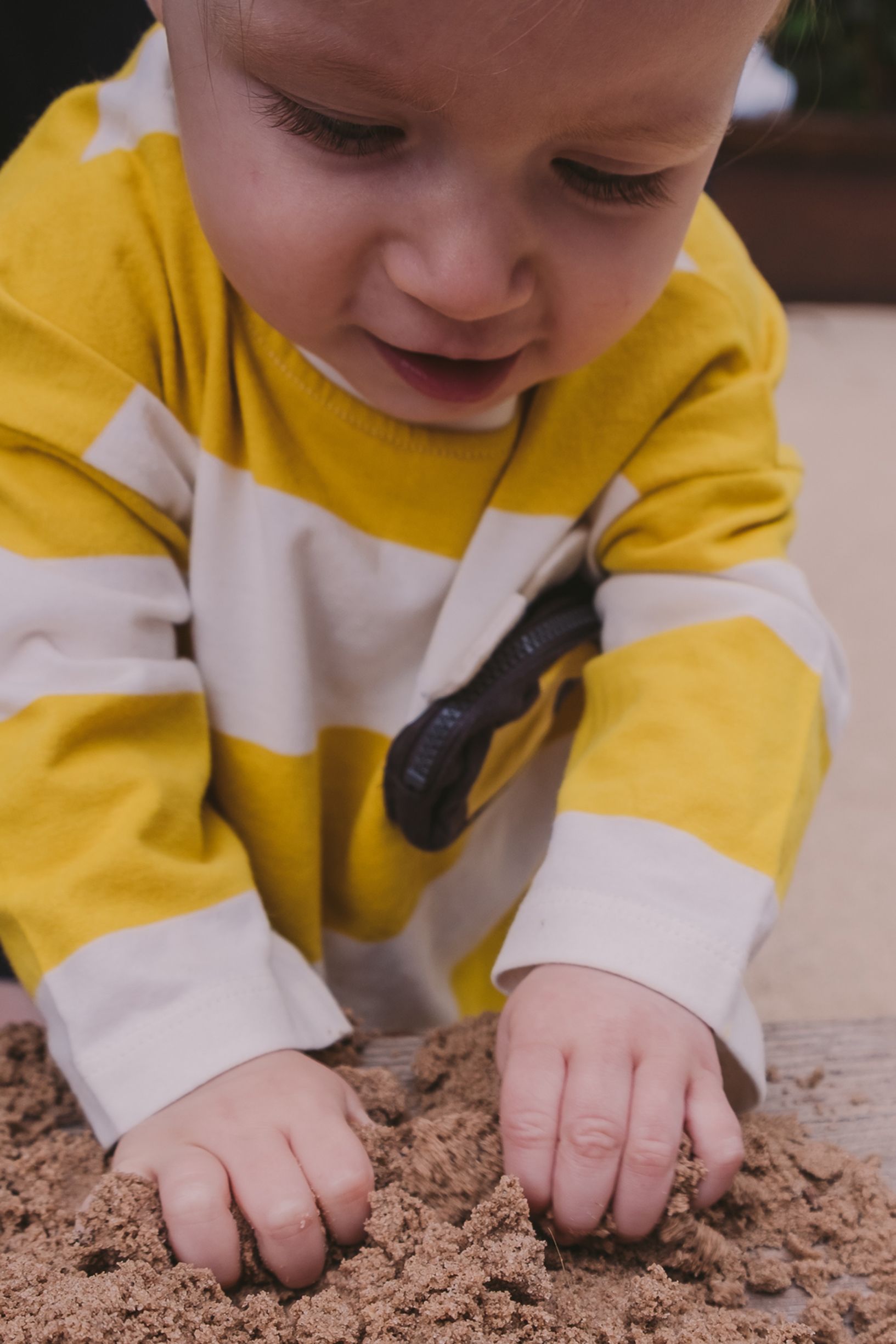 A baby in yellow and white clothing explores dirt or sand with their hands.