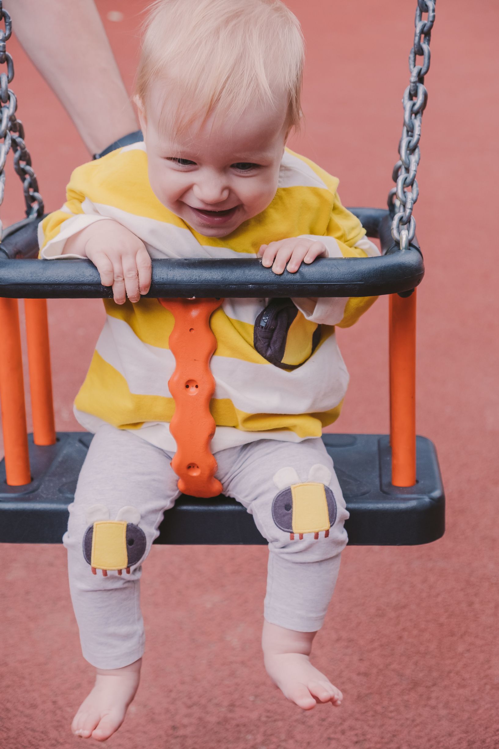 A small child wearing a yellow shirt and gray pants smiles while sitting in an orange and black playground swing.